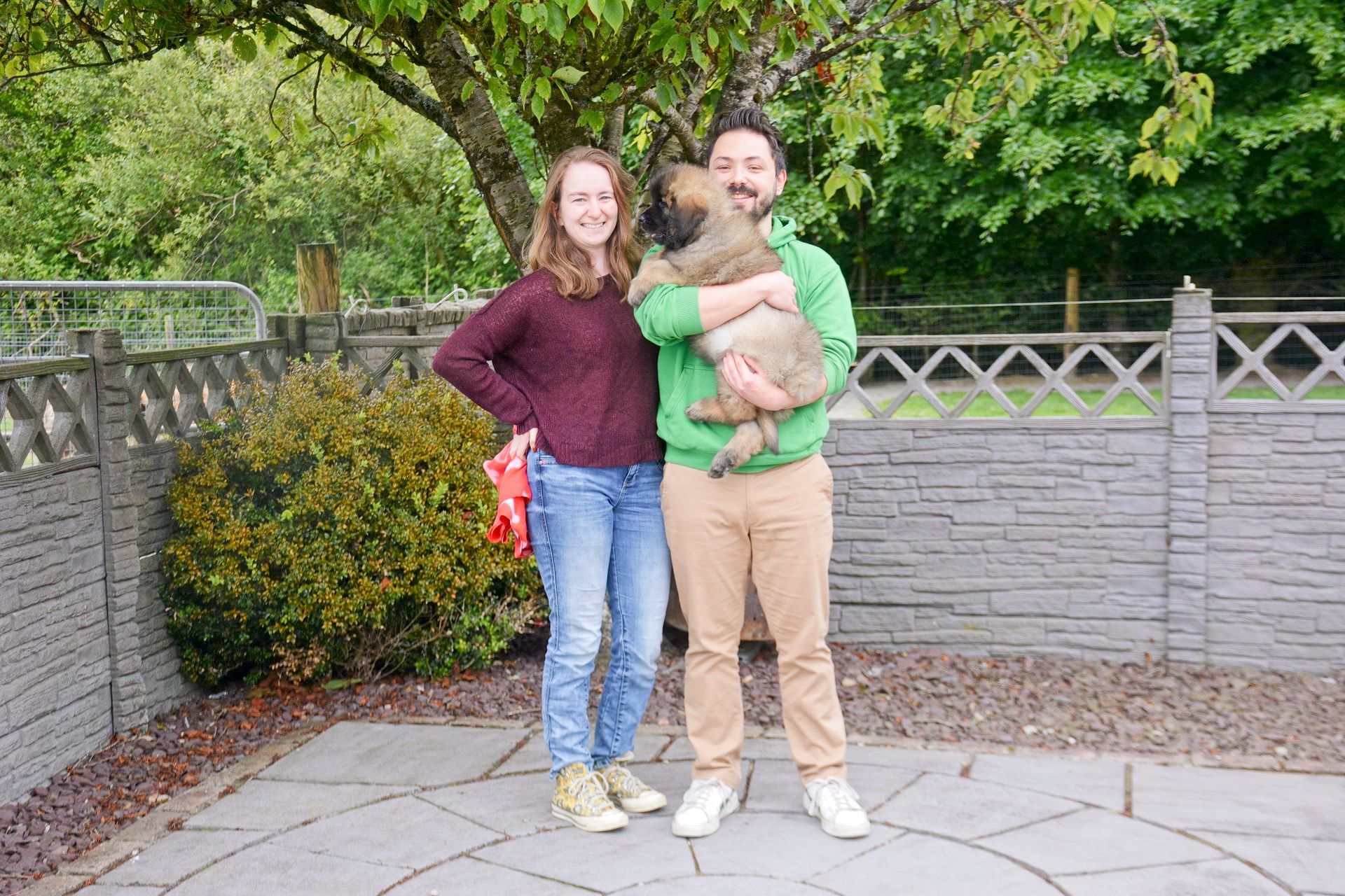 Two people standing outdoors by a gray wall, holding a small dog and smiling at the camera.