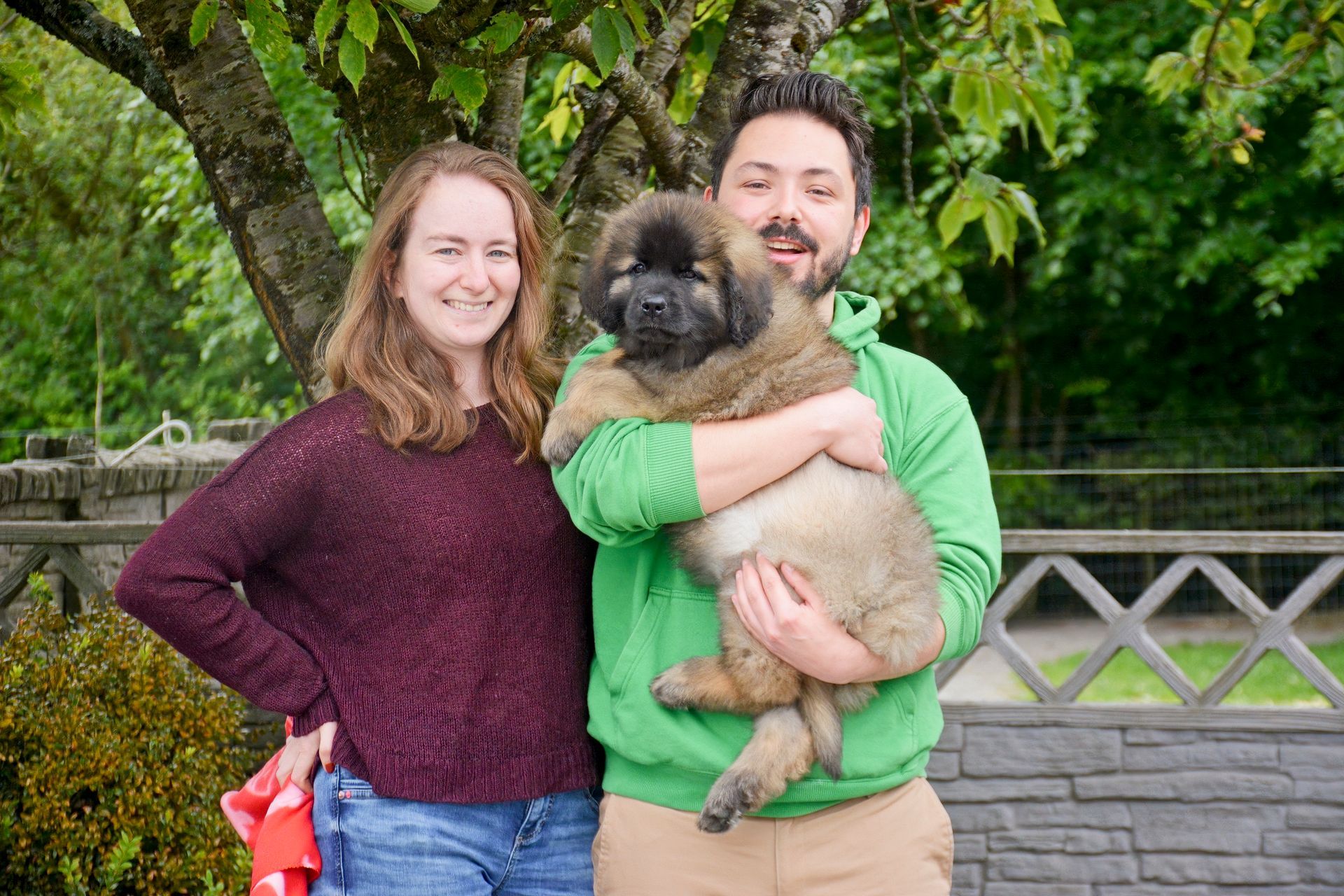 Two people outdoors, smiling while holding a fluffy dog in front of greenery and a stone wall