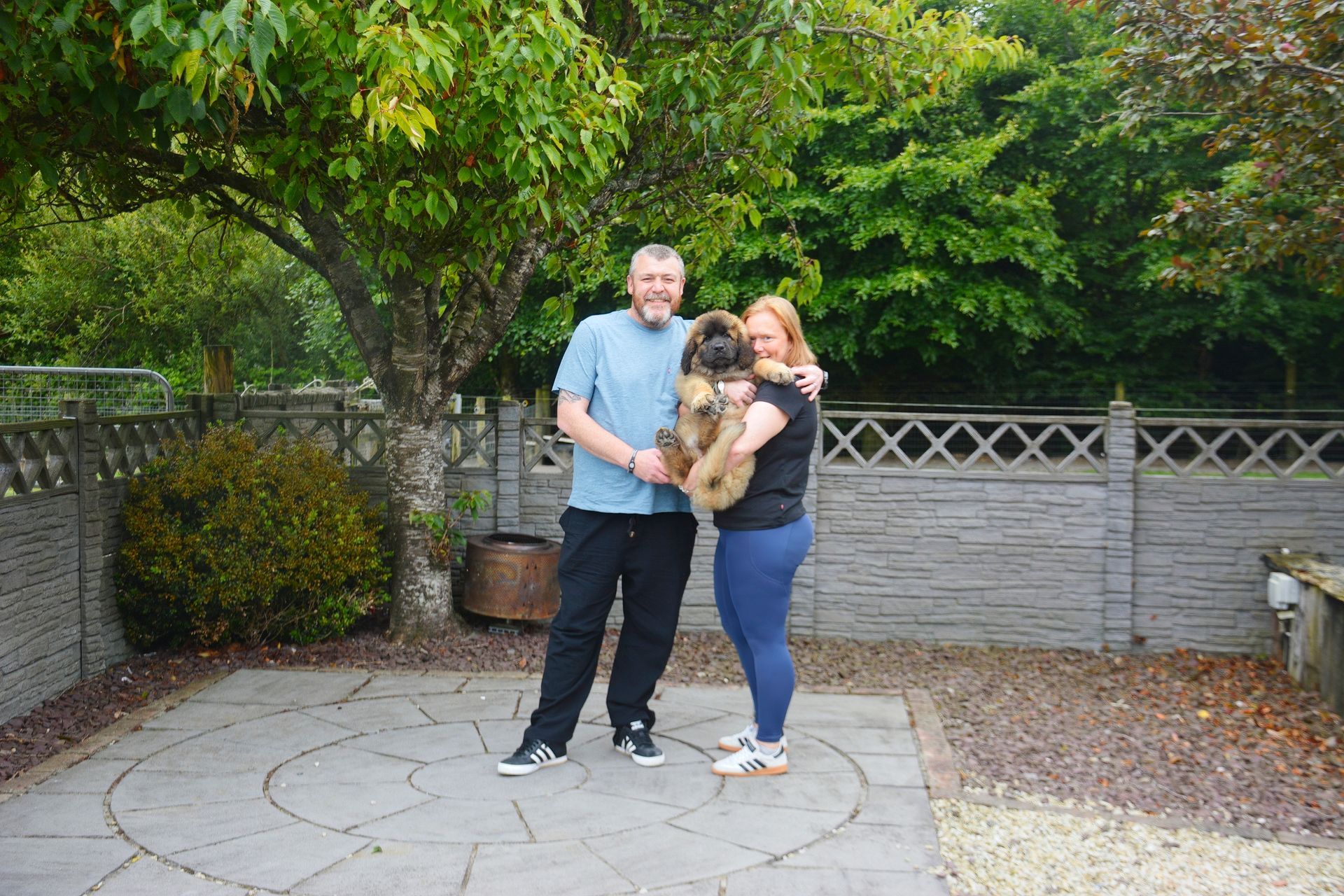 Three people standing together in a garden, holding a small dog in front of a hedge and stone wall.