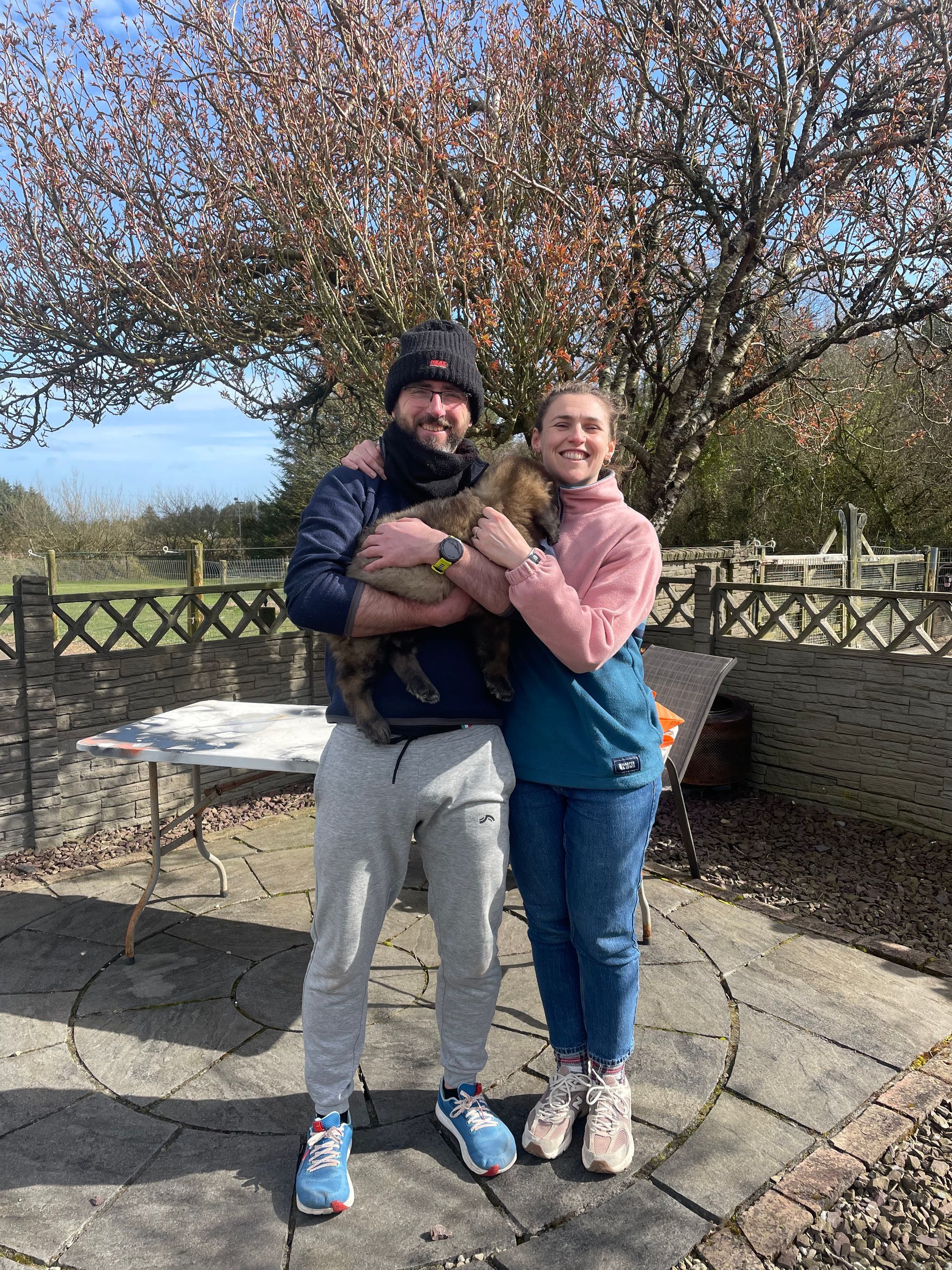 Two people smiling and holding a small dog outdoors beside a stone table under a flowering tree.