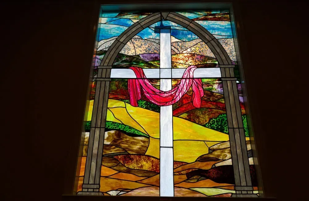 Stained-glass window with a white cross draped in red cloth over a colorful landscape background