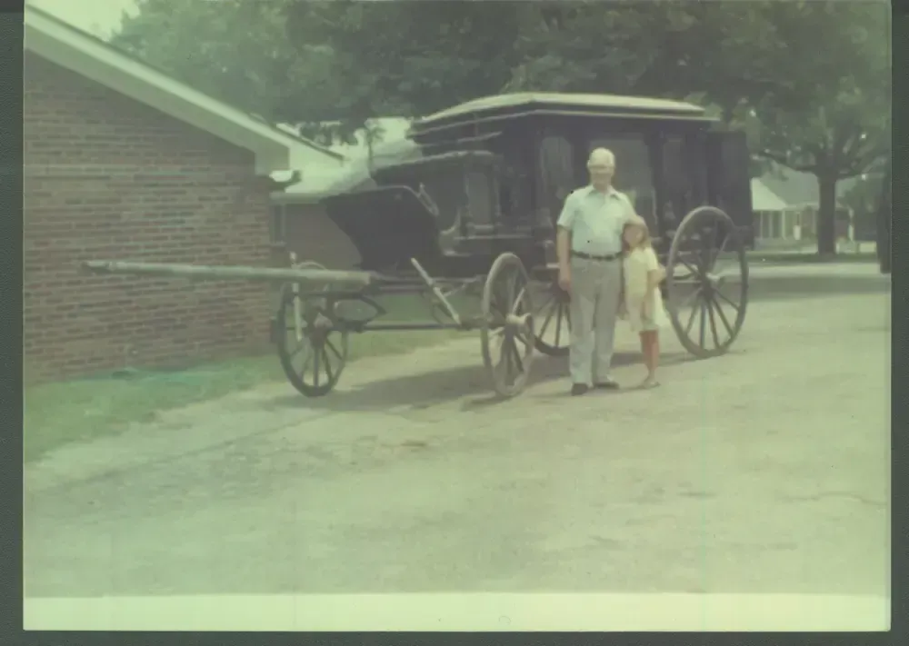 Horse-drawn hearse with two people standing beside it on a quiet street near a brick building.