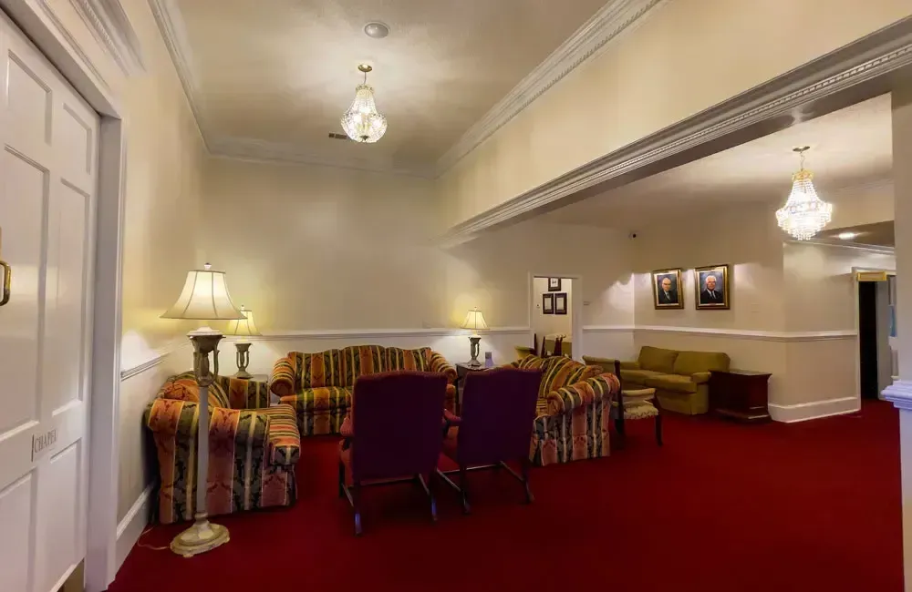 Cozy lounge with red carpet, patterned sofas, chairs, and warm lamp lighting in a white-walled room