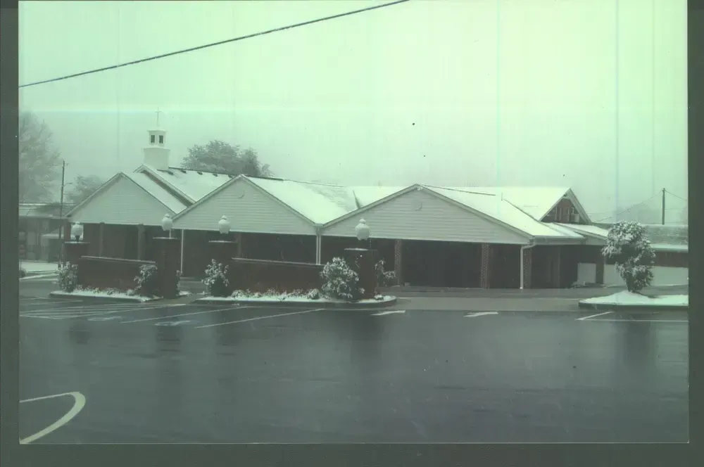 Snow-covered brick building with a white roof beside a wet road on a gray winter day