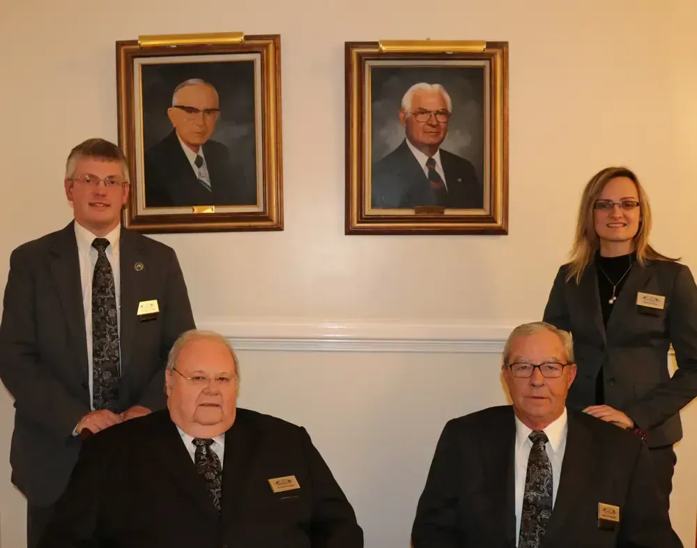 Four formally dressed people pose indoors beneath two framed portraits.