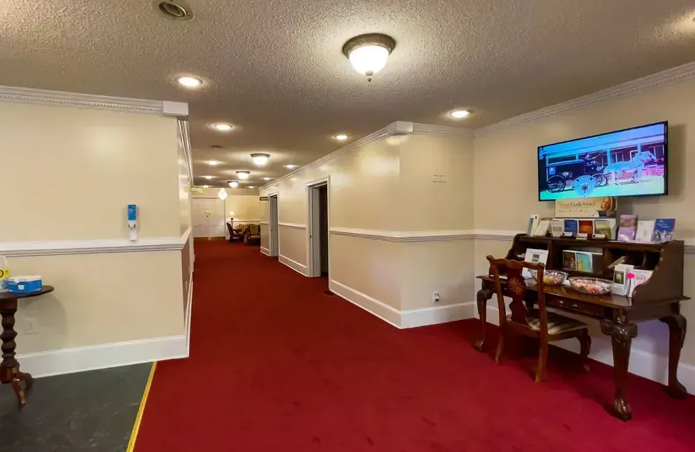 Hotel hallway with red carpet, cream walls, and a small table with a TV on the right.