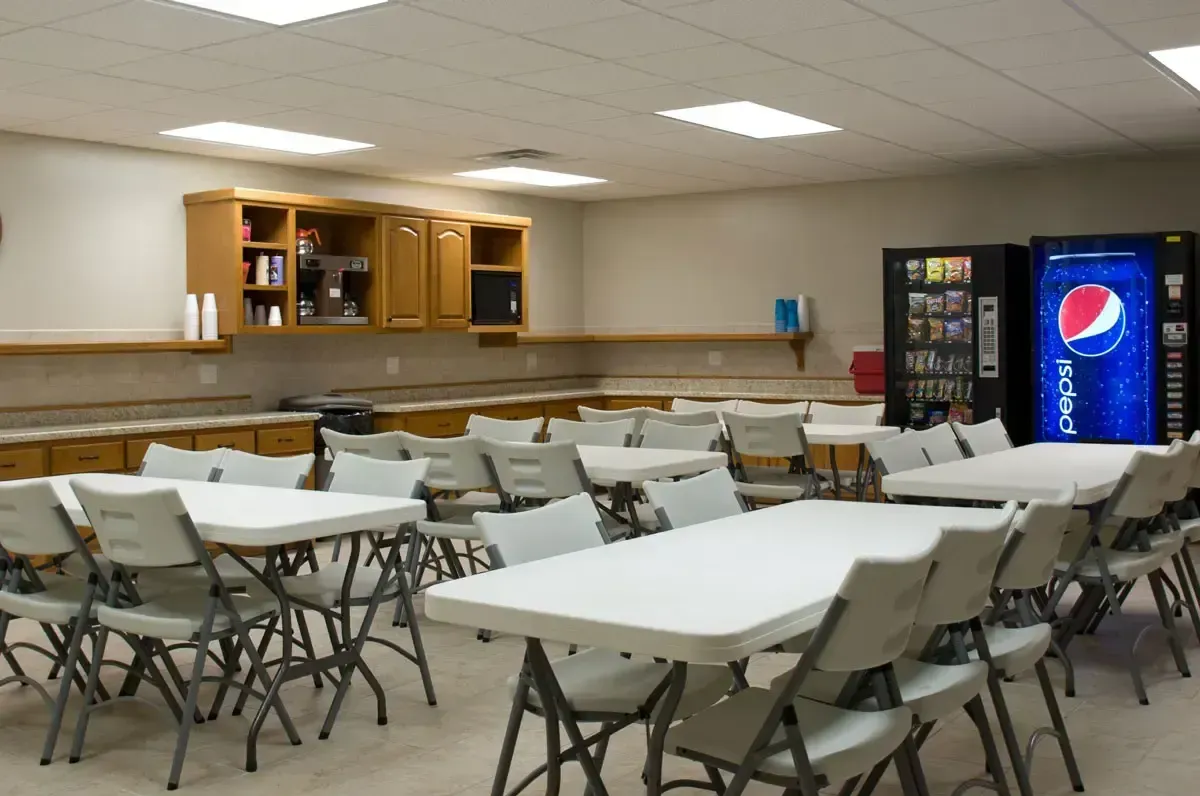 Empty cafeteria with white folding tables, chairs, wood counter, and a Pepsi vending machine.