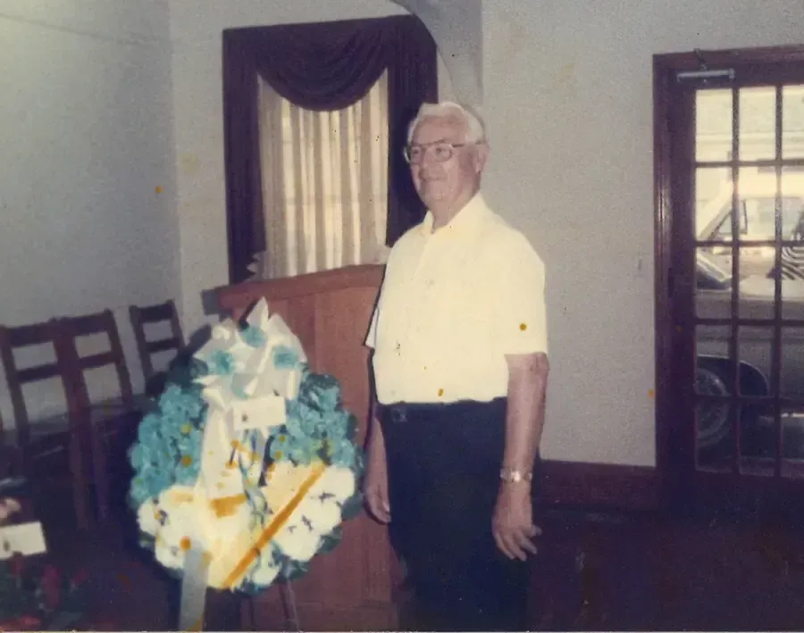 Man standing beside a blue and white floral arrangement in a room with chairs and curtained windows