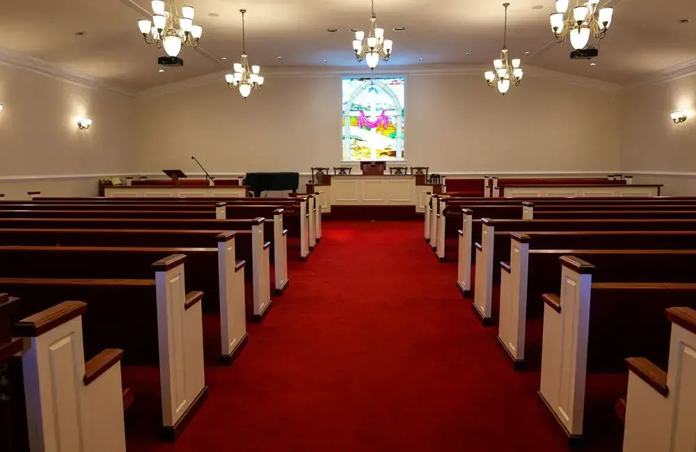Empty church interior with red carpet, wooden pews, chandeliers, and stained glass at the front