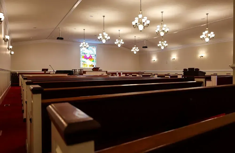 Empty church sanctuary with wooden pews, hanging lights, and a stained-glass window.