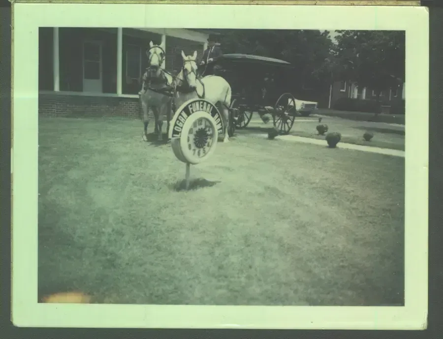 Black-and-white photo of a horse-drawn carriage on a lawn in front of a building