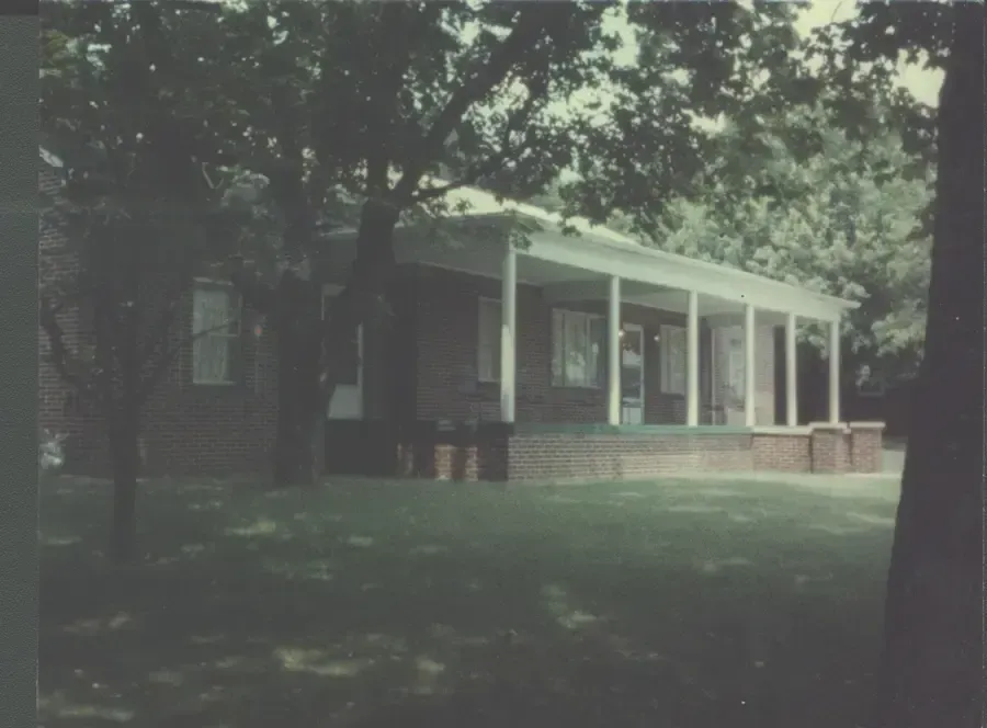 Dark house with white porch columns in a shaded yard, framed by trees.