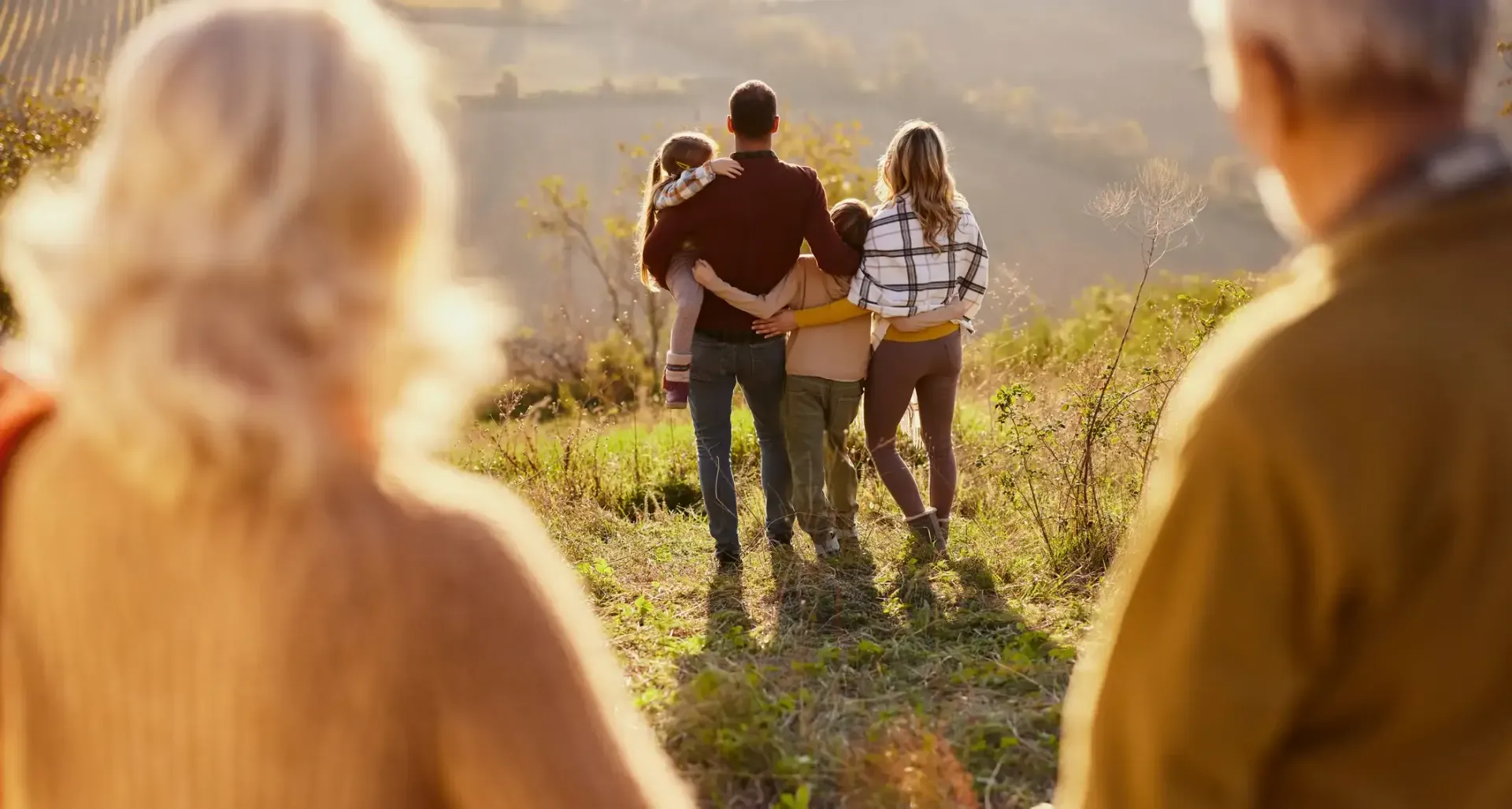 Family of five walks away from the camera, viewed by grandparents in a sunny field.