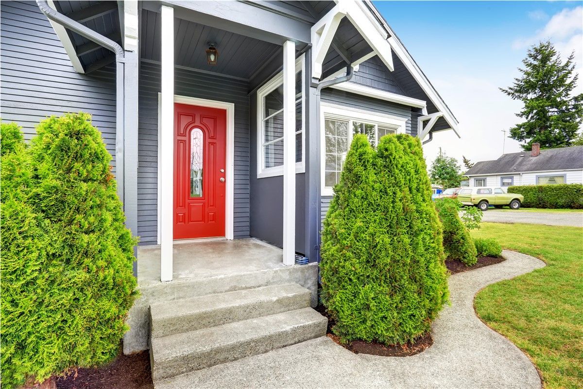 A house with a red front door and a gray exterior, featuring a small porch and green bushes.