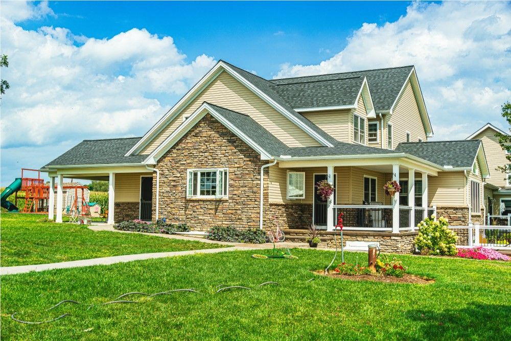 Two-story house with beige siding and stone facade; sunny day with blue sky; green lawn in front.