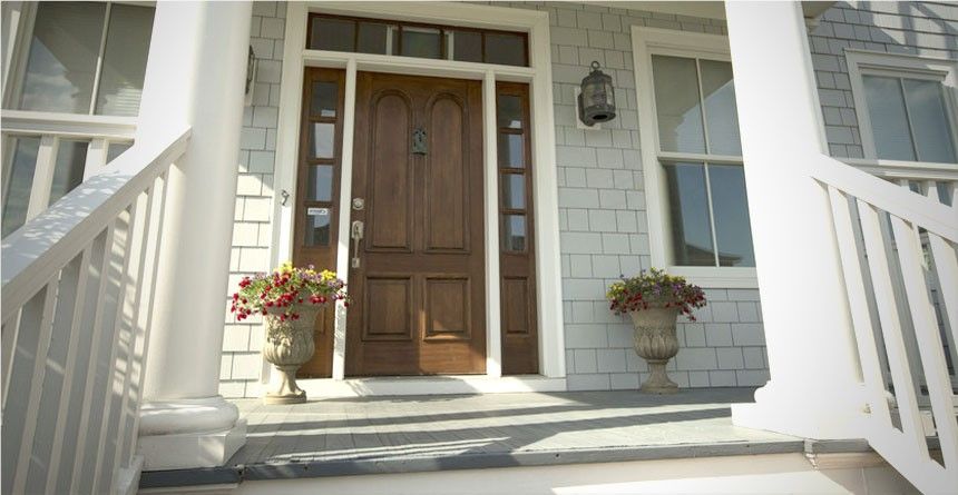 A wooden front door of a house with two planters of flowers, set on a porch with white railing.