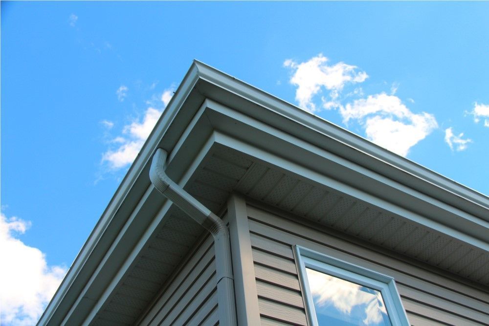 Corner of a house with light tan siding, white trim, and a gutter against a blue sky with white clouds.