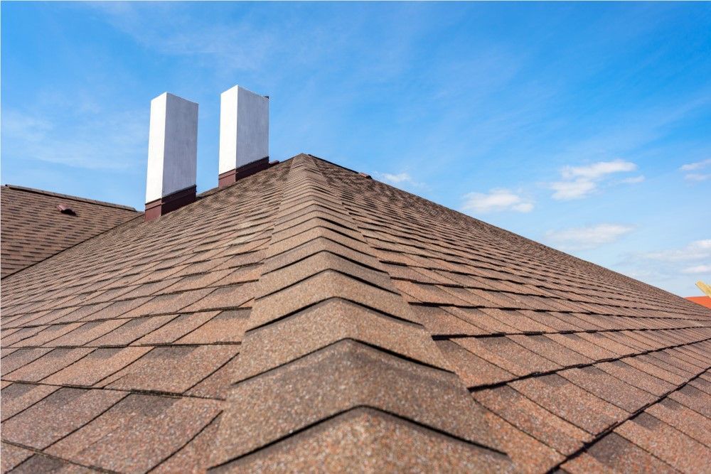 Close-up of a brown shingled roof with two white chimneys against a blue sky.