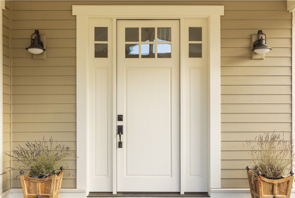 White front door with glass panel and sidelights, flanked by sconce lights and potted plants, on a beige house.