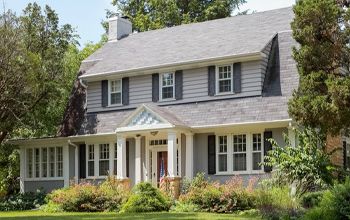 A two-story gray house with a covered porch and shutters, surrounded by trees and landscaping.