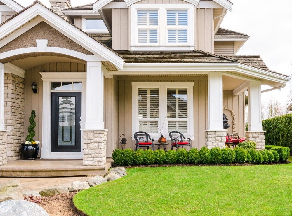 Beige two-story house with a porch and manicured lawn. Features columns, a black front door, and red chairs on the porch.