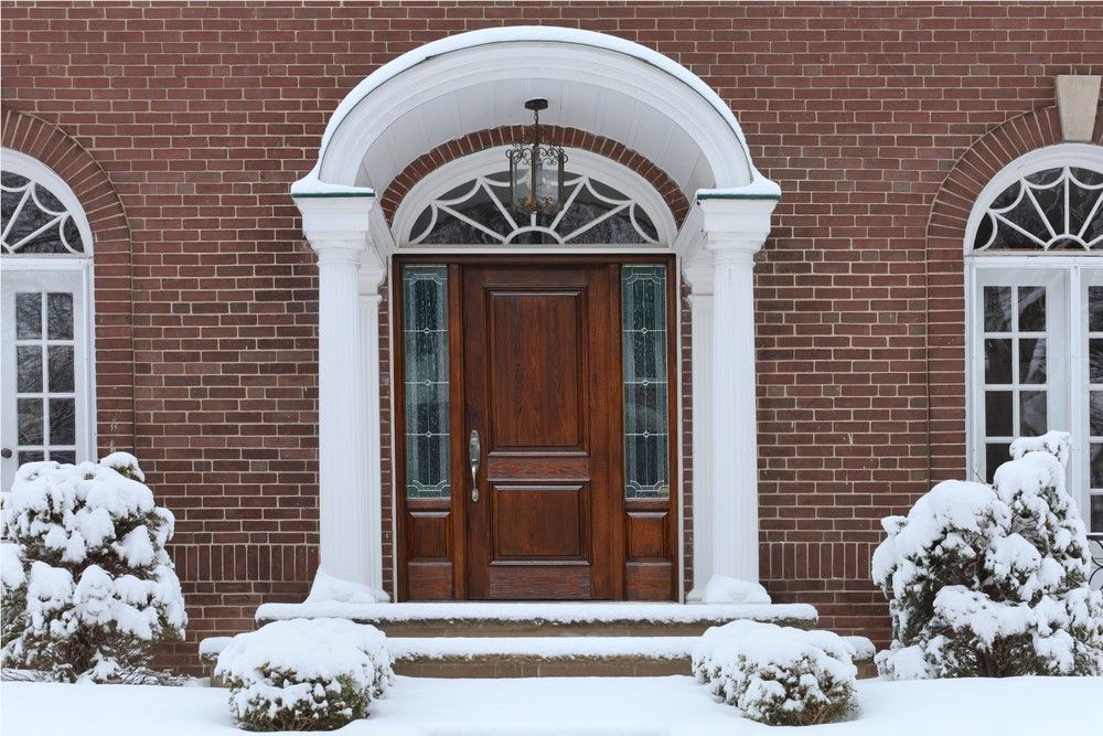 A wooden door with an arched doorway, white pillars, and a red brick exterior in a snowy winter scene.