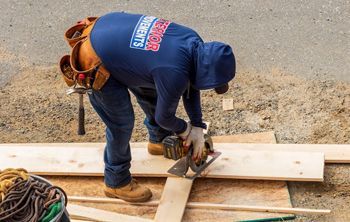 Construction worker in a blue hoodie and work boots cuts wood with a saw outdoors. He wears a tool belt and gloves.