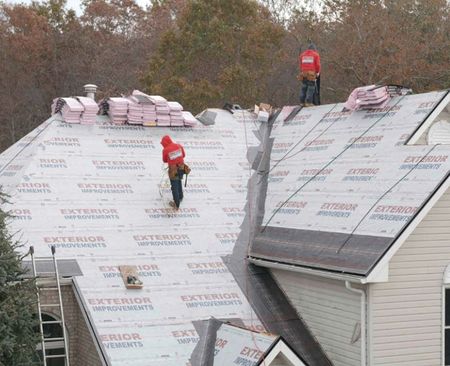 Roofers in red jackets working on a house roof, laying down materials and shingles on a cloudy day.