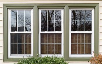 Three vertical, white-framed windows with green trim, reflecting bare trees and featuring brown blinds