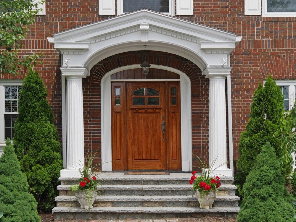 Brick house entryway with wooden door, white columns, and steps, flanked by evergreen trees and flower pots.
