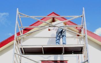 Man on a scaffolding painting the white facade of a house, with a red roof, against a blue sky.