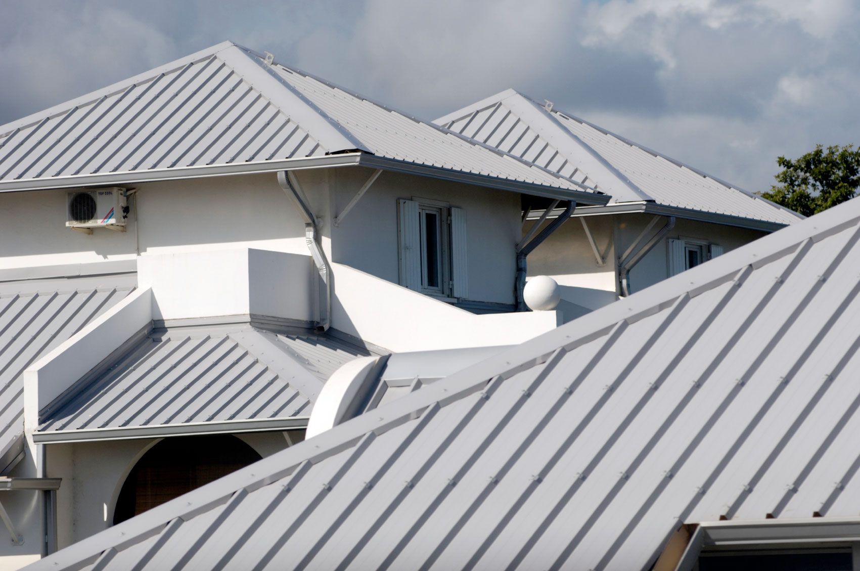 White building with multiple angled, light gray metal roofs under a cloudy sky.