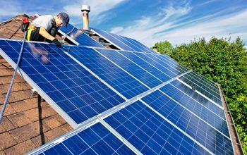 Roofer installing solar panels on a brown shingled roof against a blue sky. A person is working on the panels.