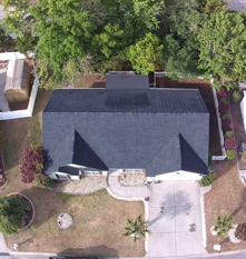Aerial view of a house with a dark roof, driveway, and front yard surrounded by trees.