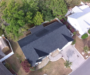 Aerial view of a dark-roofed house beside a driveway, with trees and a neighboring white-roofed home nearby.