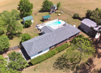 Aerial view of a rural property with a long gray-roofed house, swimming pool, trees, and small outbuildings.