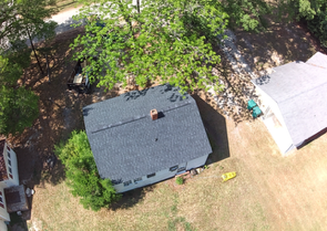 Aerial view of a house with a dark roof, surrounded by trees, lawn, and a driveway with a white vehicle.