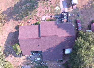 Aerial view of a house with a brown roof, surrounded by trees, yard debris, and a parked red vehicle.