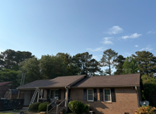 Single-story brick house with brown roof under a clear blue sky, surrounded by trees
