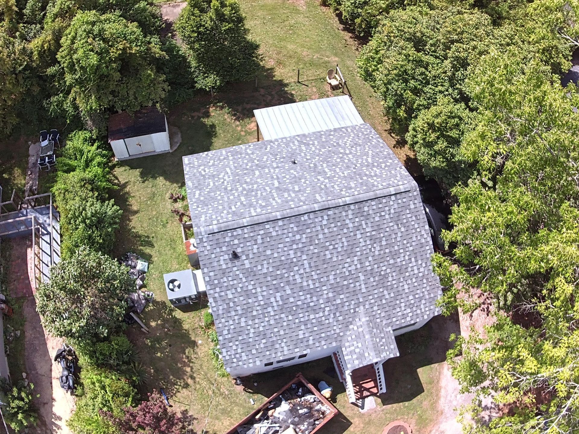 An aerial view of a house with a new grey shingled roof, surrounded by trees, a lawn, and a dumpster in the yard.