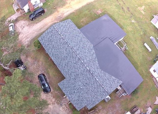 An aerial view of a house with a grey shingled roof, next to a larger flat-roofed section, surrounded by grass and cars.