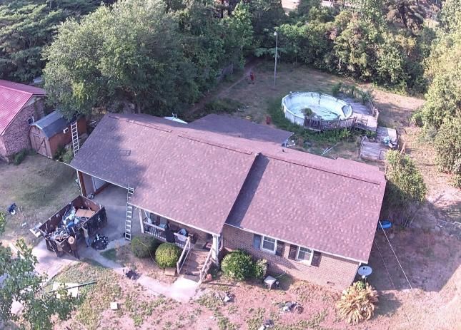 Aerial view of a house with a brown roof, red shed, and backyard trees beside a round pool