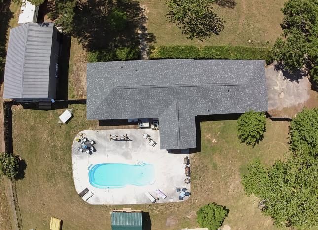 Aerial view of a backyard with a house, patio, and swimming pool surrounded by lawn and trees