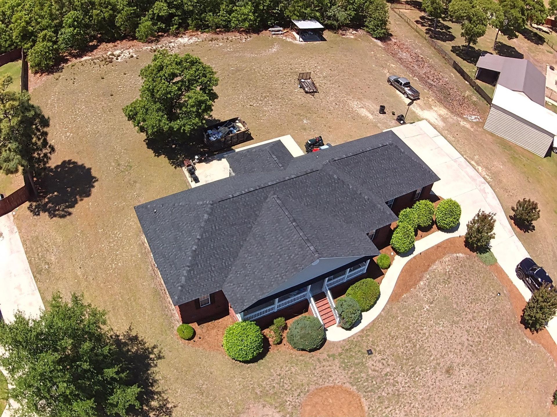 Aerial view of a house with a dark roof, driveway, trees, and a large dry lawn.