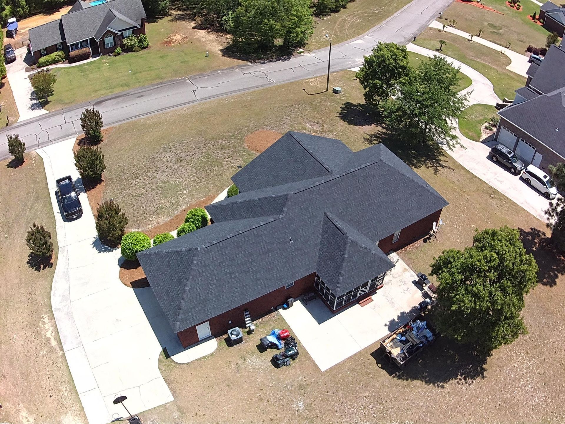 Aerial view of a dark-roofed suburban house with driveway, trees, and neighboring homes.