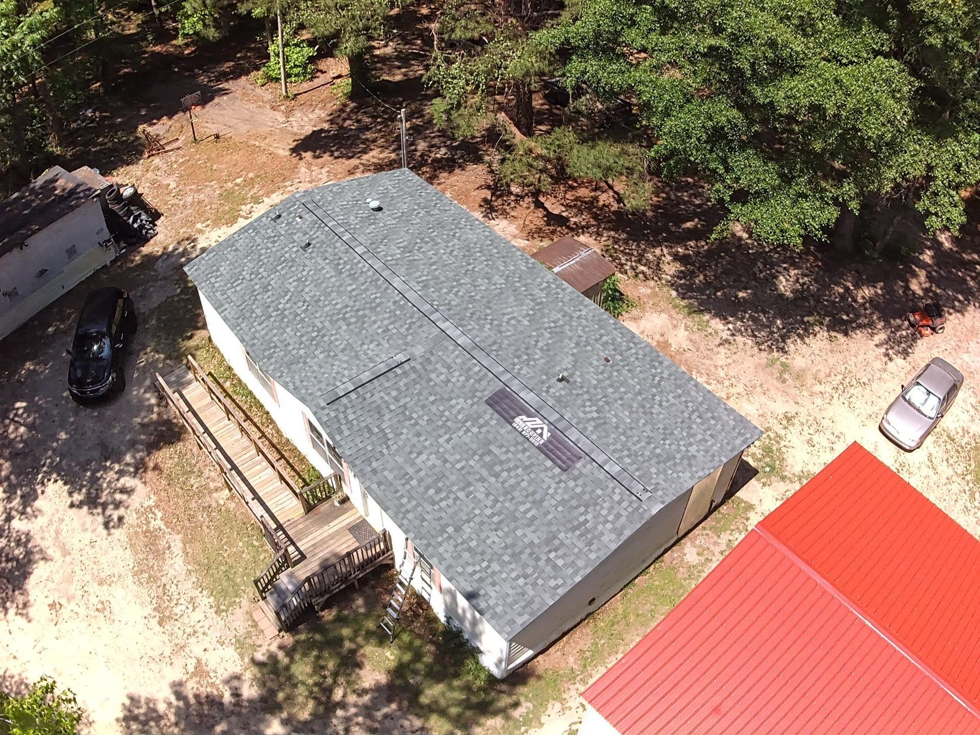 Aerial view of a gray-roofed building beside a red-roofed structure, surrounded by trees and dirt paths