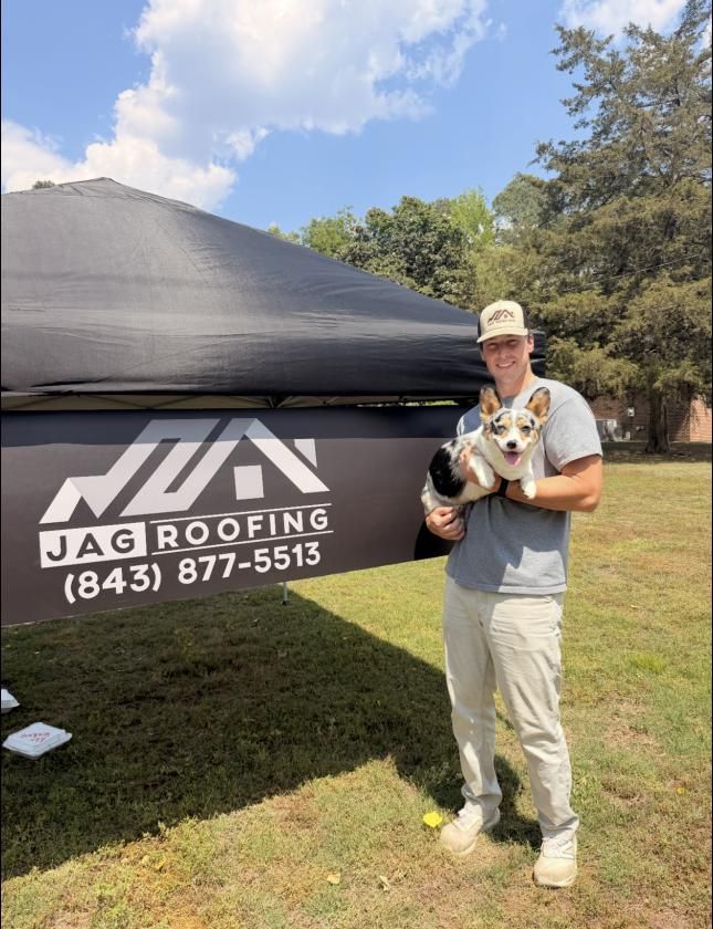 Man holding a small dog beside a JAG Roofing tent on a sunny lawn