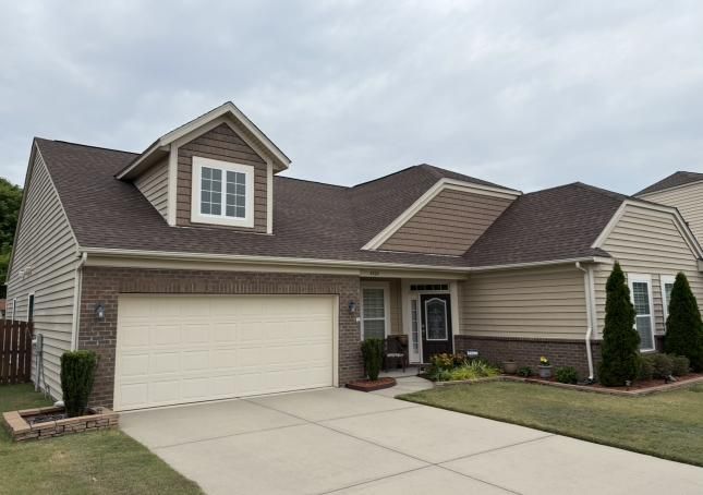 Suburban tan house with attached garage, concrete driveway, and a small front lawn under a cloudy sky