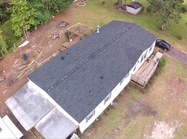 Aerial view of a long white house with a dark roof beside a wooded yard and parked car.