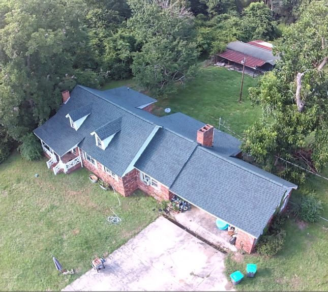 Aerial view of a brick house with a gray roof, driveway, and trees surrounding it.